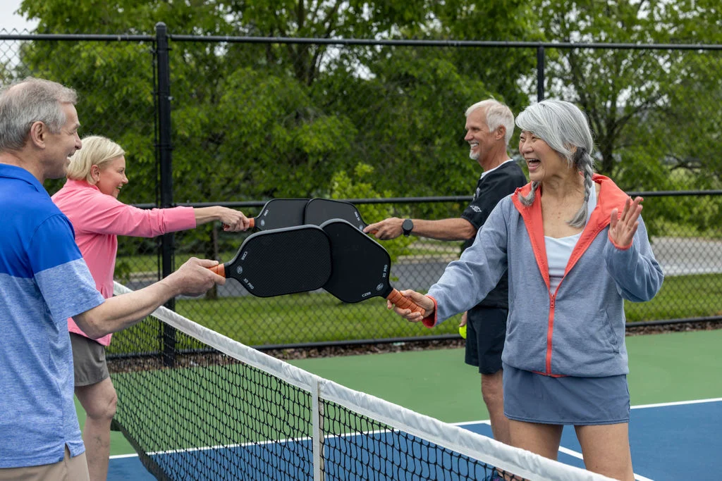 Older adults holding paddles and laughing together at the net of an outdoor court