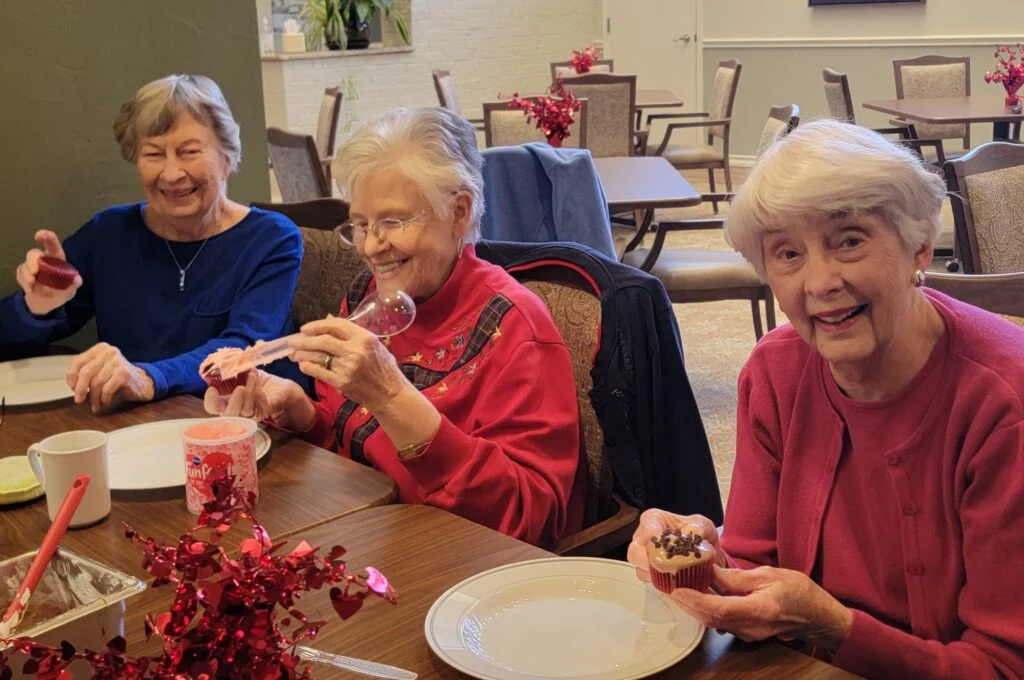Three smiling older women seated at a table with plates and red flower decorations