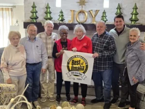 Group of smiling older adults posing together in a holiday-decorated room, holding up a small award banner