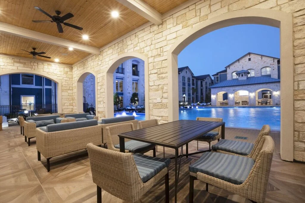 Covered stone patio with lounge seating and a dining table, with an arched opening overlooking a pool and apartment buildings