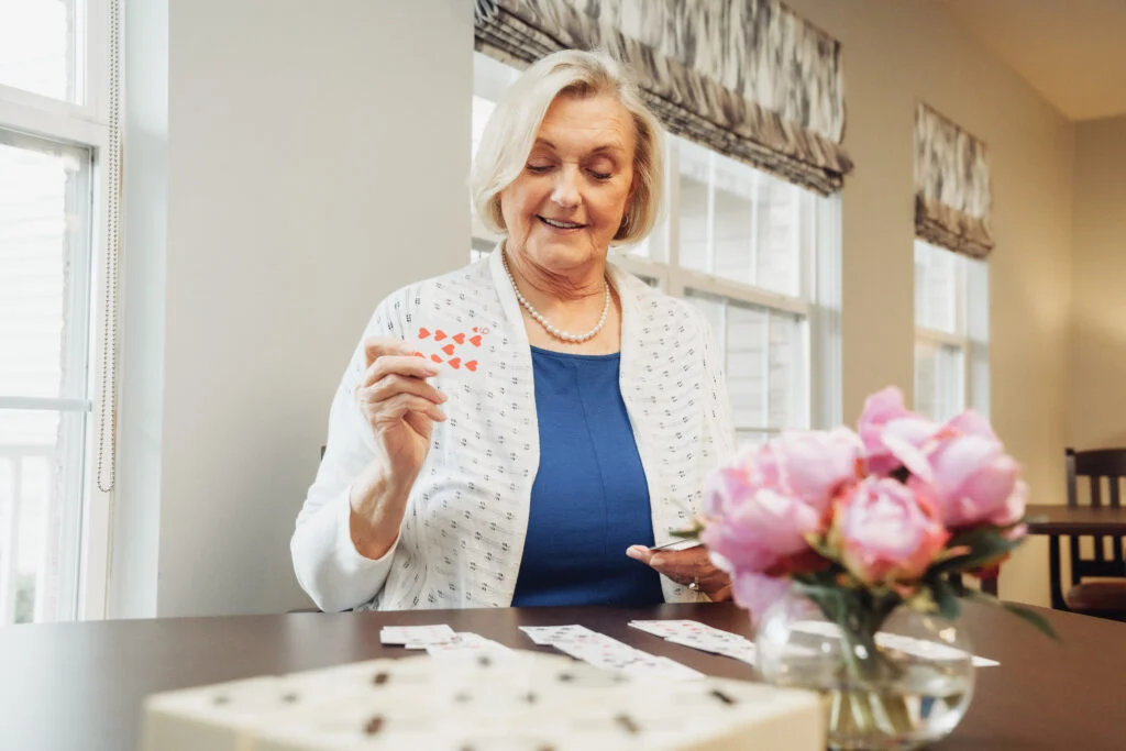 Older woman in a white cardigan holding a card at a table with a vase of pink flowers