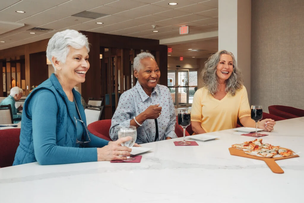 Three smiling older women chatting at a table with glasses of wine and a board of pizza slices