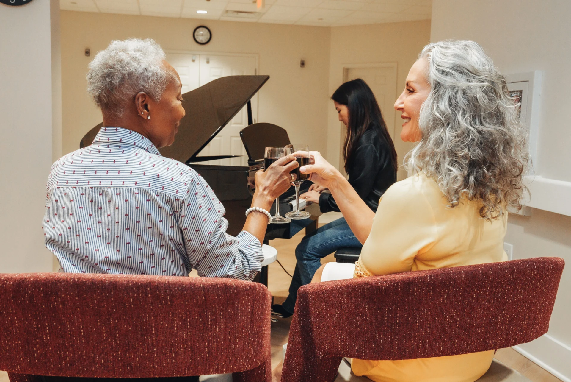 Group of older adults seated in red armchairs near a grand piano, talking and smiling