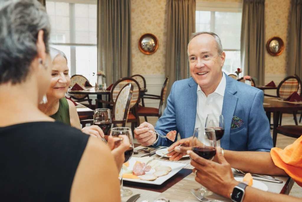 Smiling older man in a blue blazer holding a glass of red wine, seated at a dining table with friends