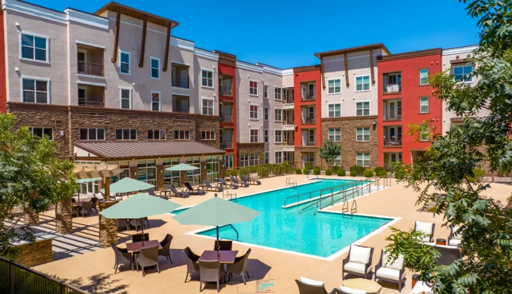 Courtyard swimming pool with patio tables and teal umbrellas, surrounded by red-accented apartment buildings