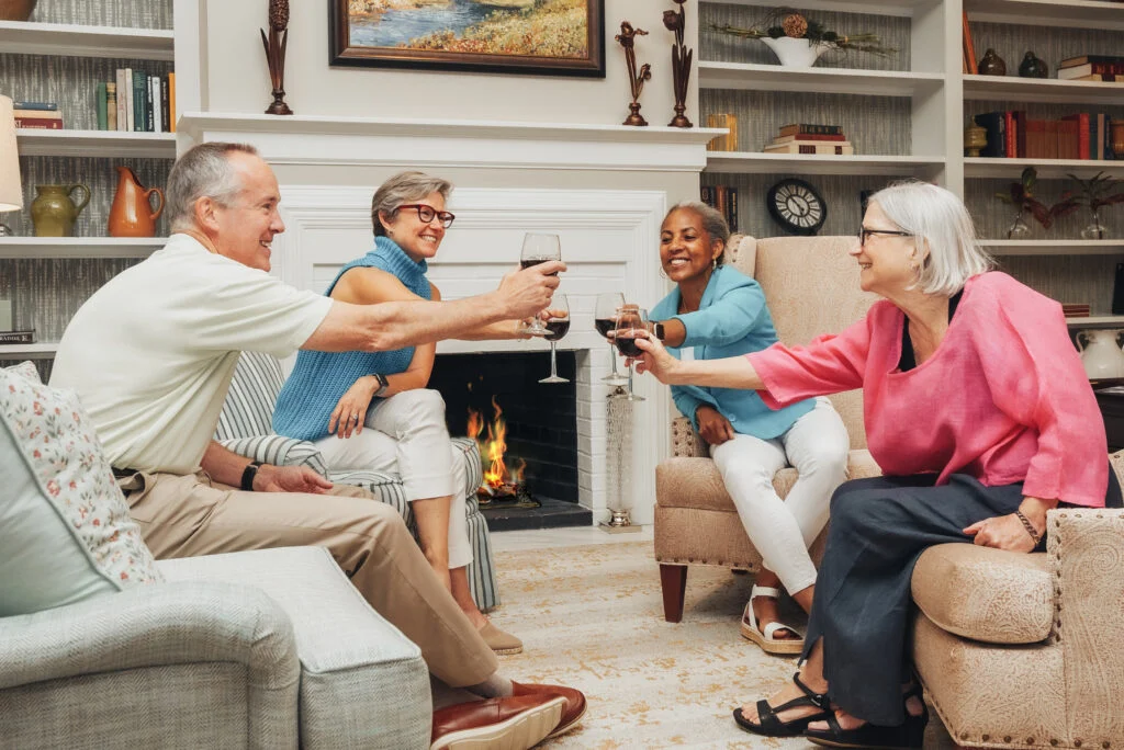 Group of older adults seated by a fireplace, clinking glasses of wine together