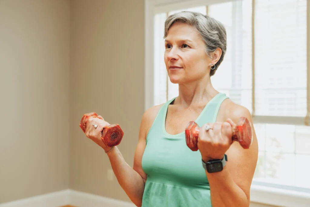 Older woman in a teal tank top lifting red hand weights in a bright room