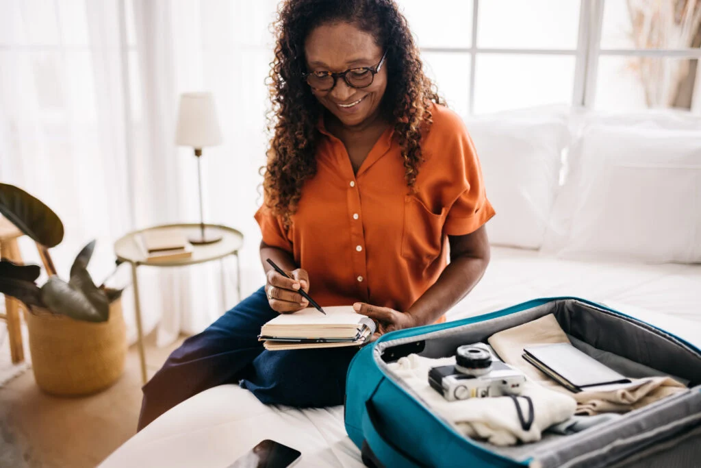 Woman in an orange blouse writing a list beside an open suitcase packed with clothes