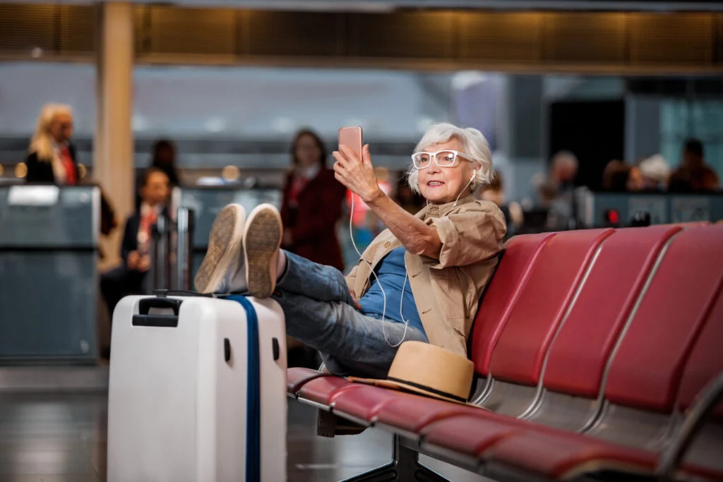 Older woman in a sun hat lounging on a red airport bench beside a rolling suitcase, holding up her phone