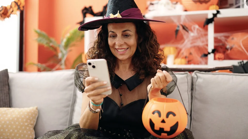 Woman in a witch costume holding a jack-o'-lantern bucket and looking at her phone on a sofa
