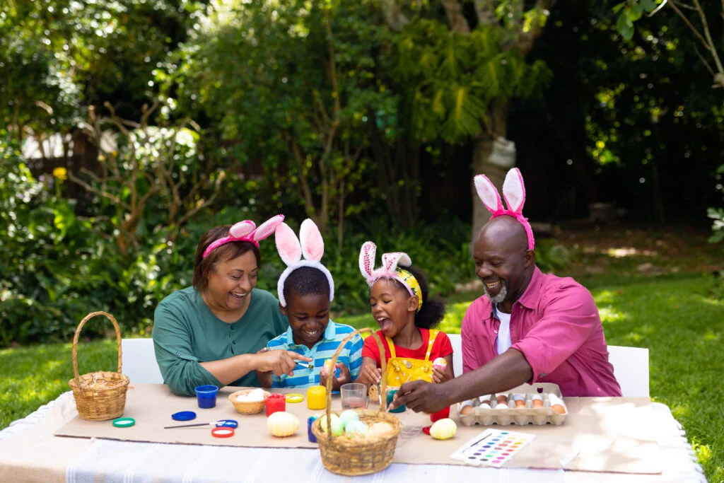 Adults and children wearing bunny-ear headbands decorating eggs at an outdoor table