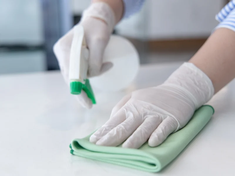 Gloved hands wiping down a white counter with a green cloth and a spray bottle