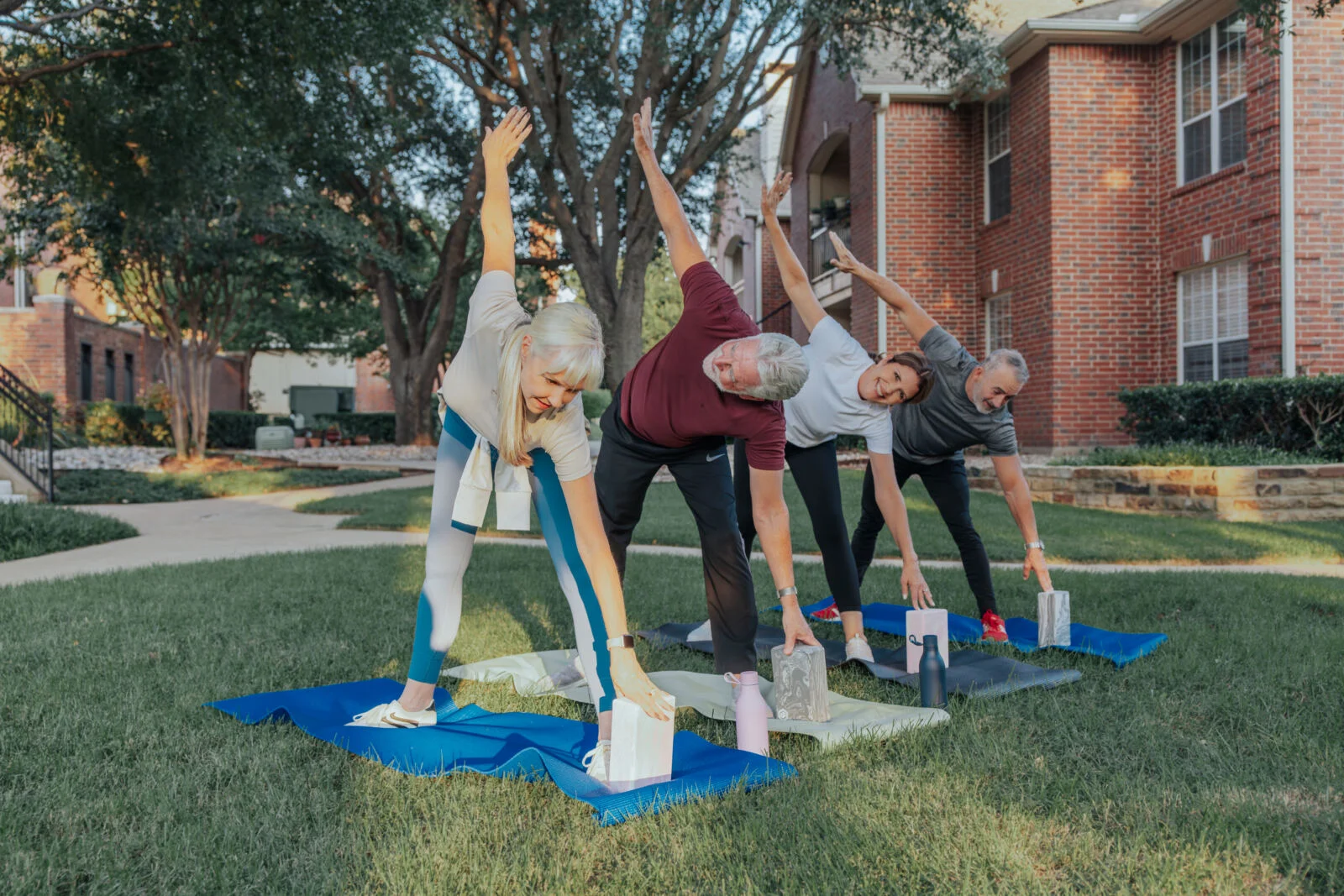 Group of older adults stretching on blue yoga mats on a lawn outside brick apartment buildings