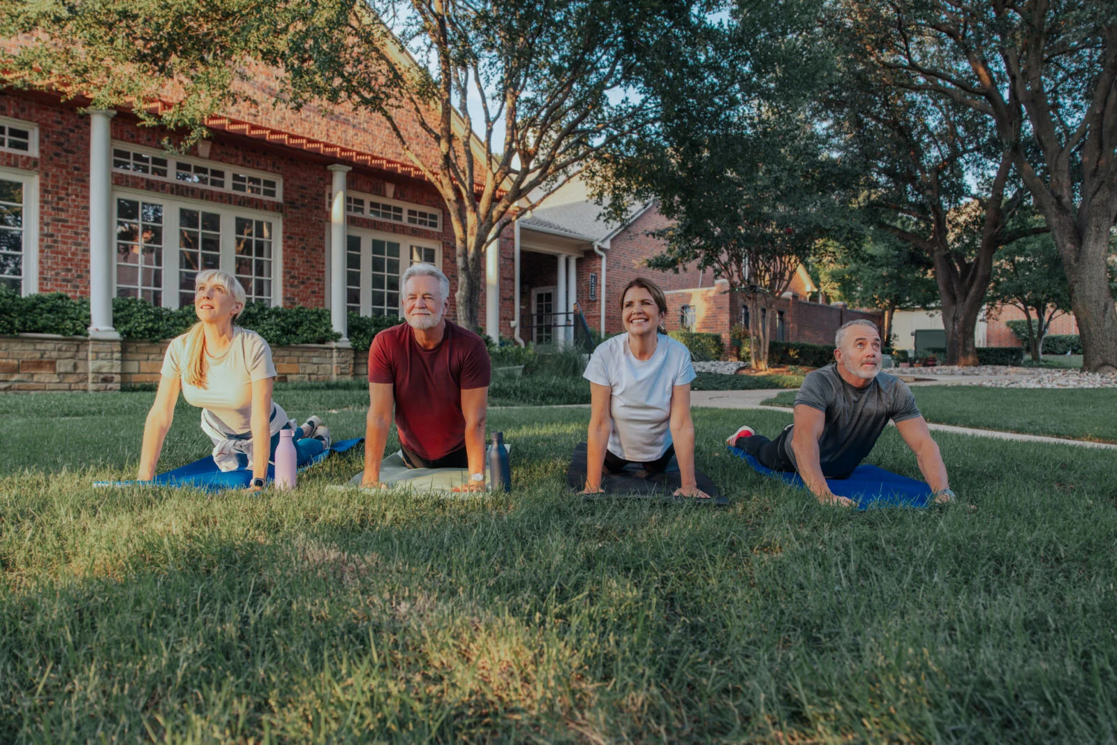 Four older adults doing yoga on mats on a lawn in front of brick buildings