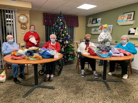 Group of residents in face masks seated at round tables in a decorated room with a Christmas tree, with stacks of knitted winter hats and other donations