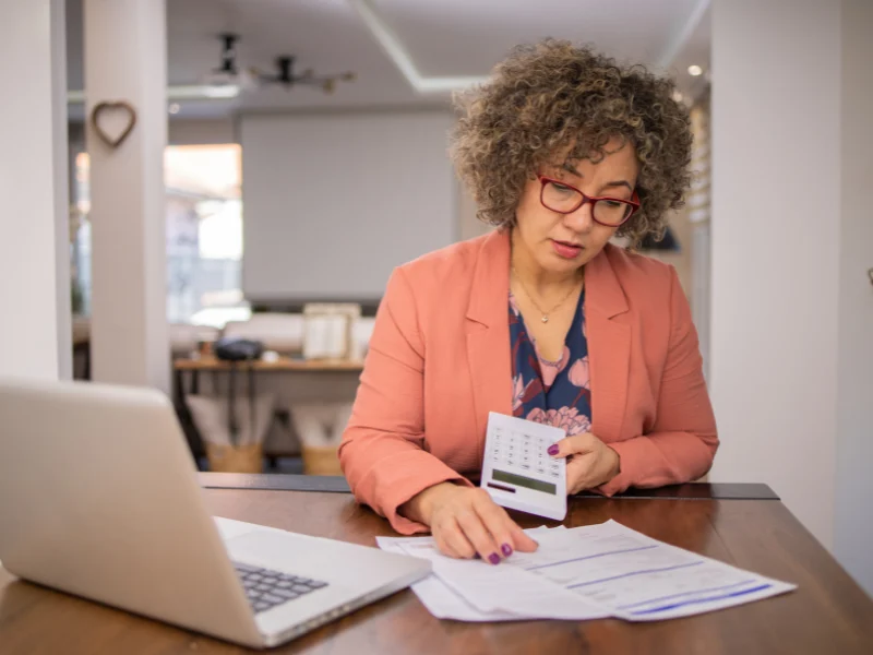 Woman in a coral blazer holding a calculator and reviewing paperwork at a desk with a laptop