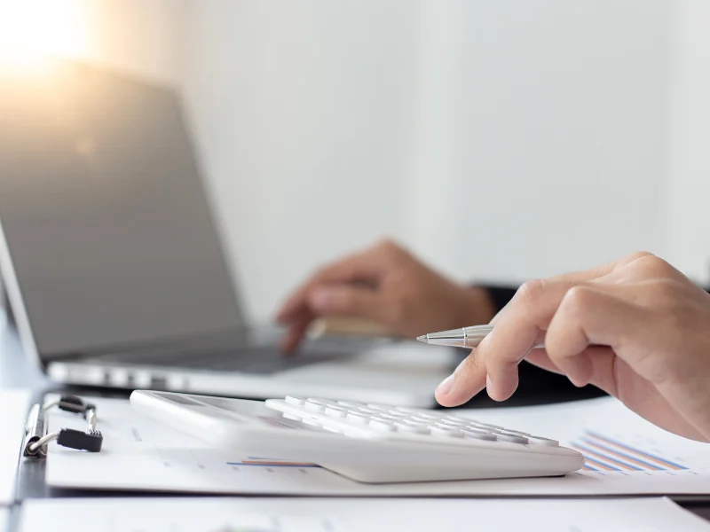 Close view of a hand using a calculator beside a laptop and paperwork