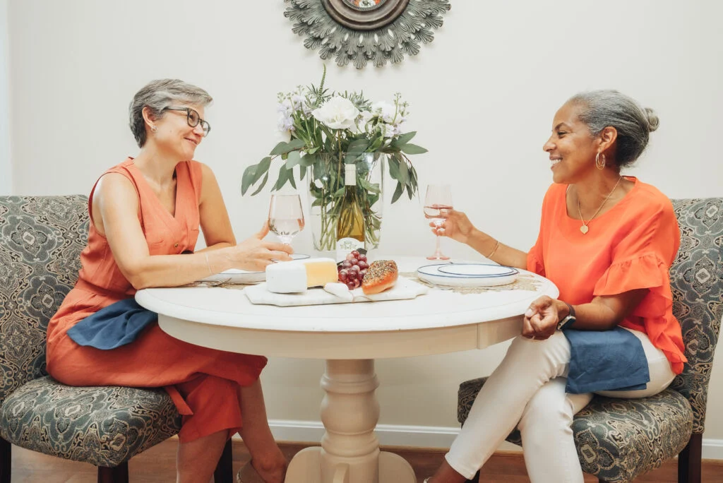Two older women in orange tops smiling and sharing wine at a round table with a flower arrangement