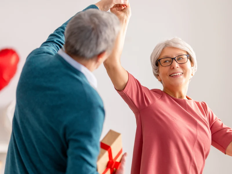 Older couple dancing together, the woman smiling in a red top, with a gift and a heart balloon nearby