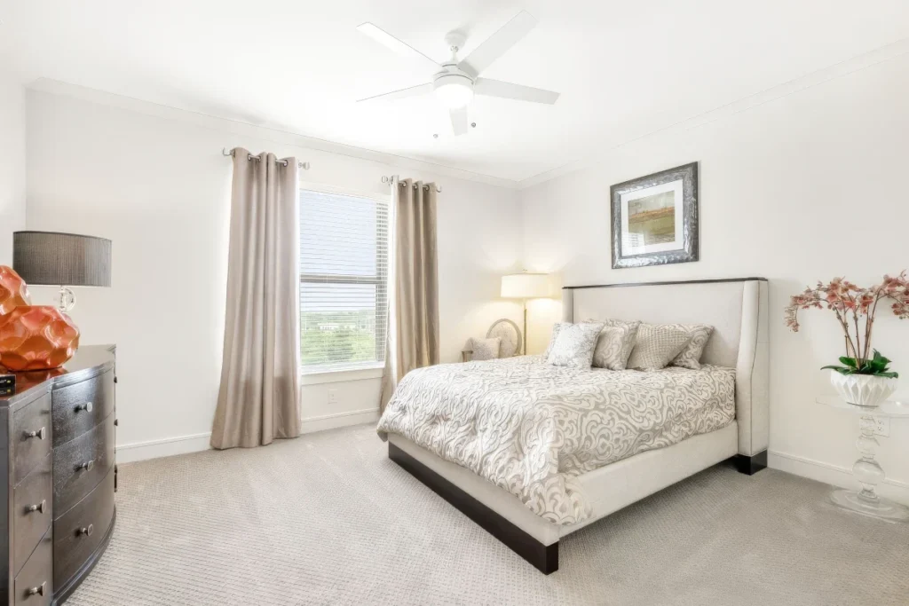 Bedroom with a white upholstered bed, beige curtains, a ceiling fan and a dresser with an orchid