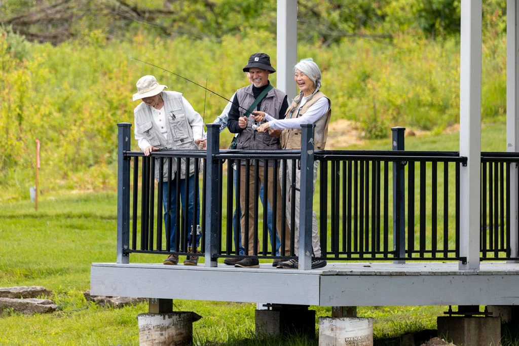 Three older adults standing on a wooden deck overlooking green fields