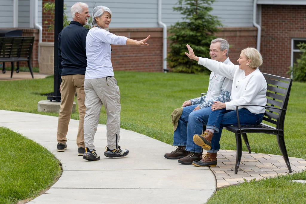 Older adults on a paved walkway, two seated on a bench and two standing, chatting and gesturing outdoors