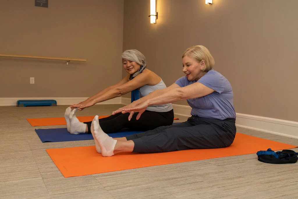 Two older women stretching while seated on colorful yoga mats in an exercise room
