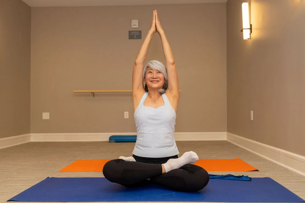 Older woman sitting cross-legged on a yoga mat with her hands raised overhead in a studio