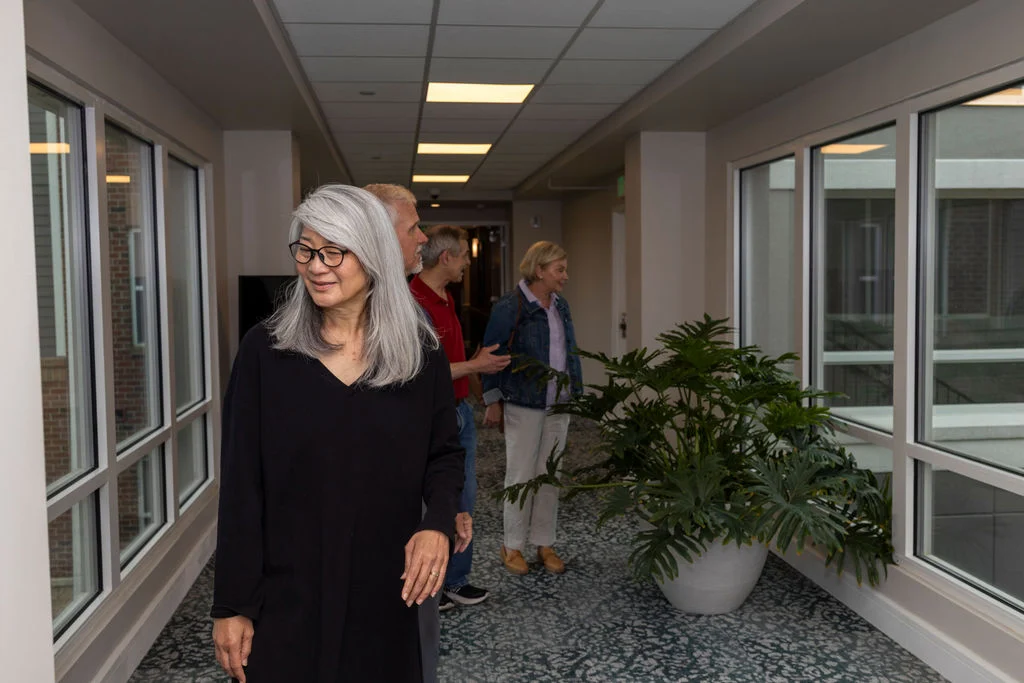 Woman in a black dress standing in a bright hallway with residents walking behind her