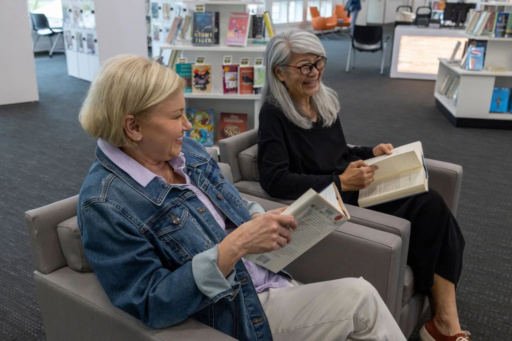 Two older women sitting in armchairs and reading books in a bright library
