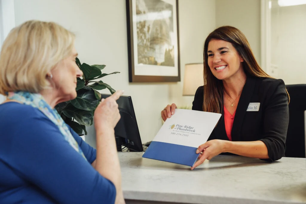 Smiling staff member in a black blazer talking with an older woman across a desk, with a folder between them
