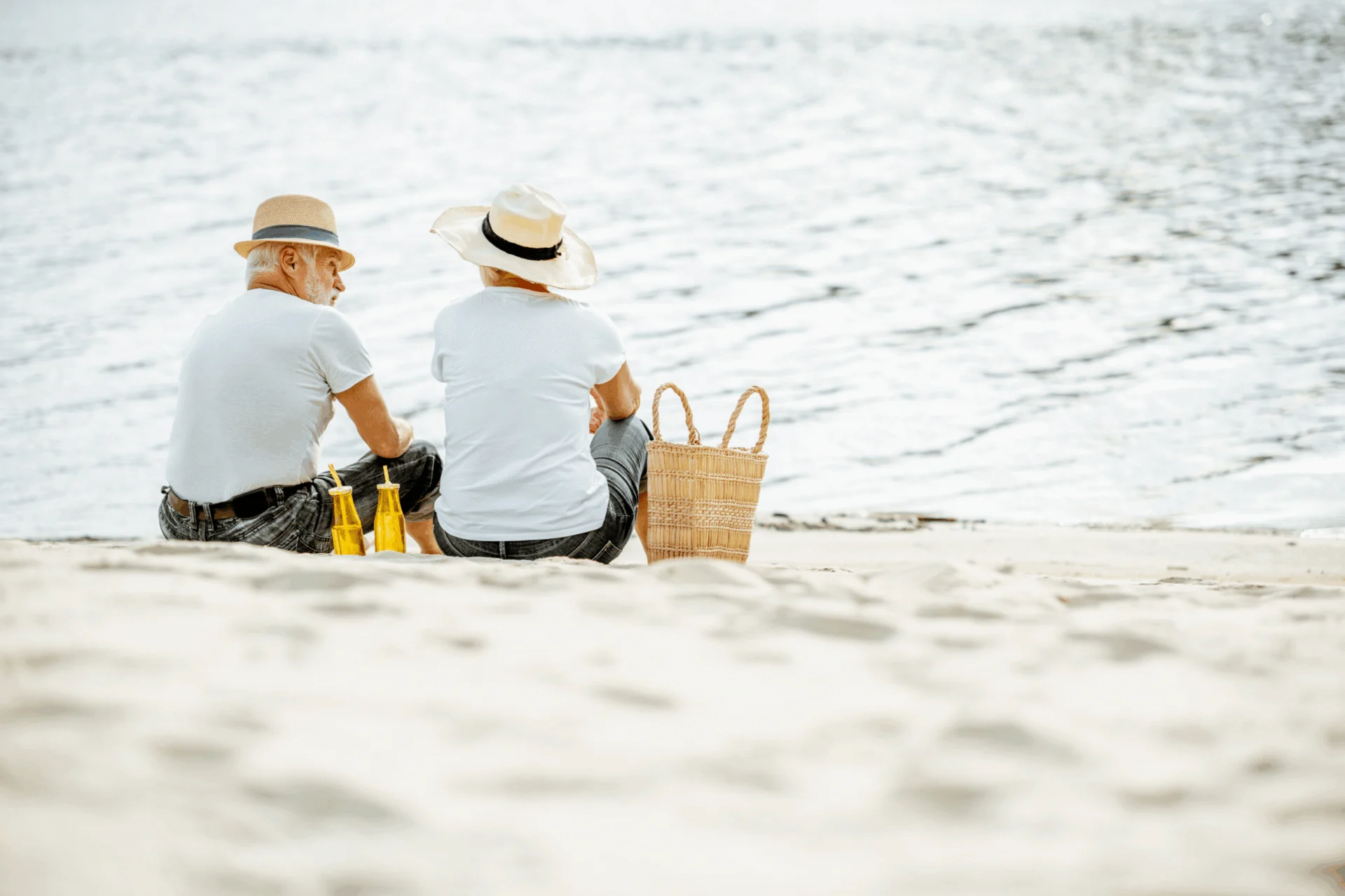 Two people in sun hats sitting on a sandy beach looking out at the water, with a picnic basket beside them
