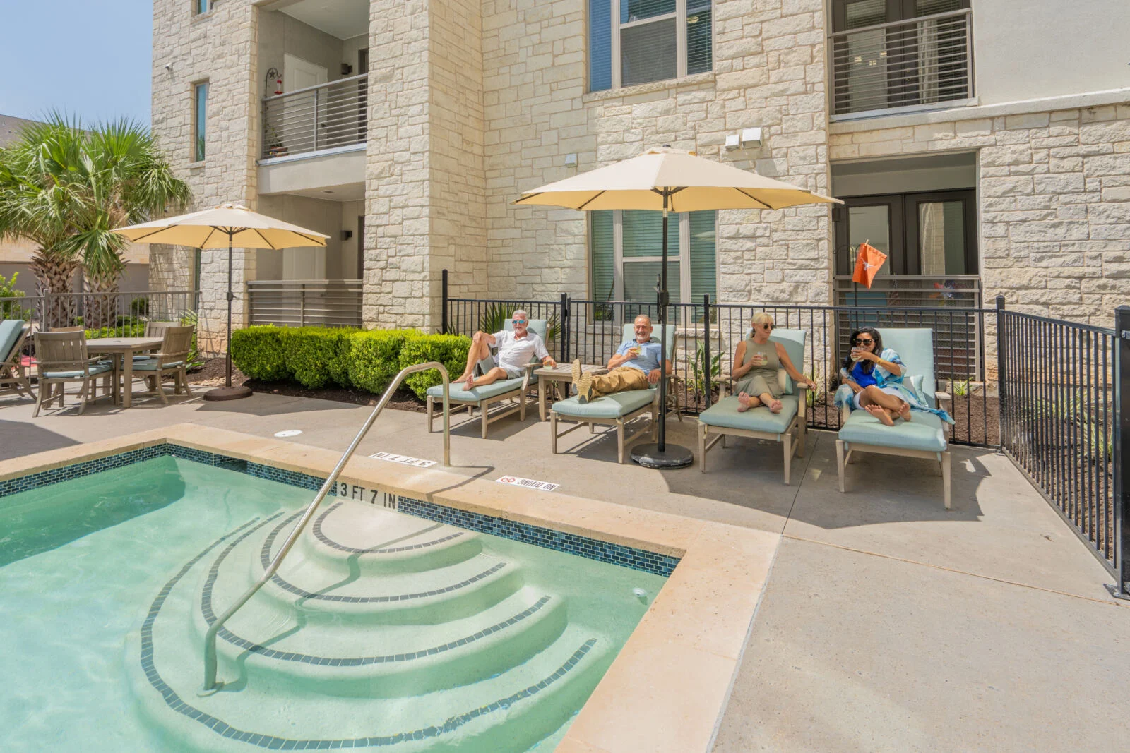 Small pool with steps and lounge chairs under umbrellas in front of a stone building, with people relaxing