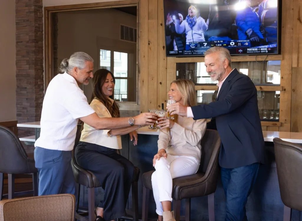 Four people toasting drinks at a bar with a TV above
