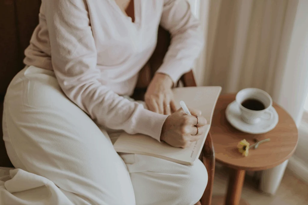 Person in cozy white clothing holding a journal, with a cup of coffee on a small wooden table beside them