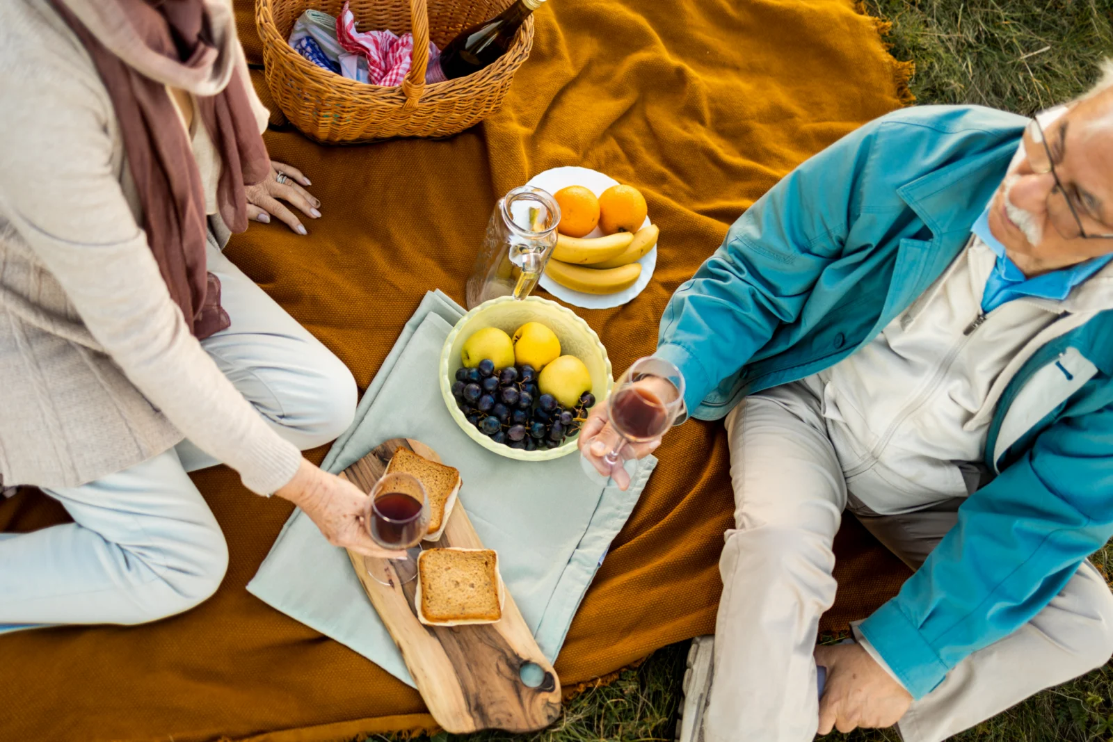 Overhead view of an older couple sharing a picnic on an orange blanket with fruit, a cheese board and wine