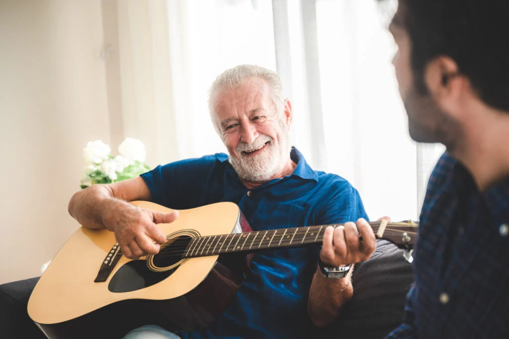 Older man smiling while playing an acoustic guitar for a younger man beside him