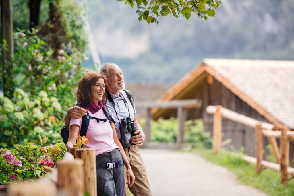 Older couple with backpacks and binoculars smiling on a leafy garden path