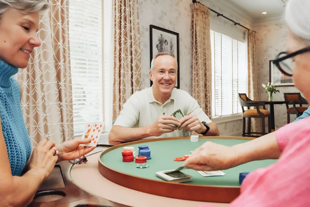 Senior man in a light shirt smiling as he plays cards at a green game table with poker chips