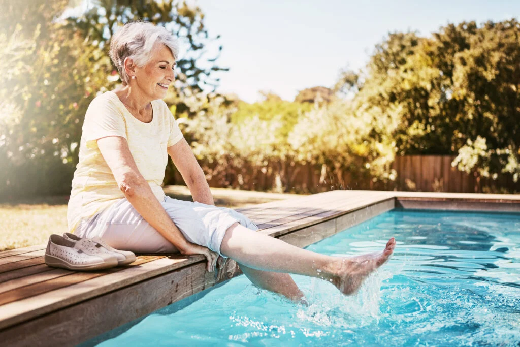 Relax, travel and senior woman by the pool while on a vacation, adventure or outdoor trip in summer. Happy, smile and elderly lady in retirement with her feet in the swimming pool at a holiday resort.