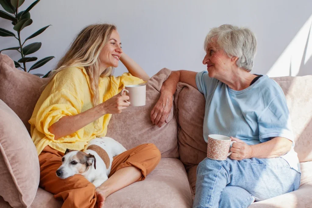 Two older women chatting with mugs on a couch with a small dog