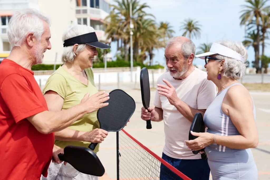Group of seniors in sun hats chatting beside a pickleball net with paddles