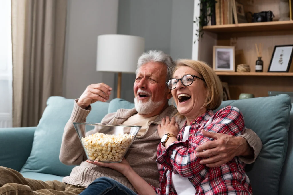 Older couple laughing together on a sofa with a bowl of popcorn