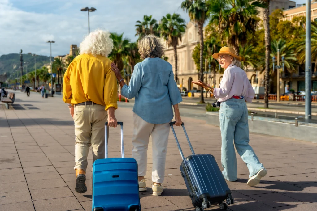 Three older travelers walking along a sunny promenade, two pulling blue rolling suitcases