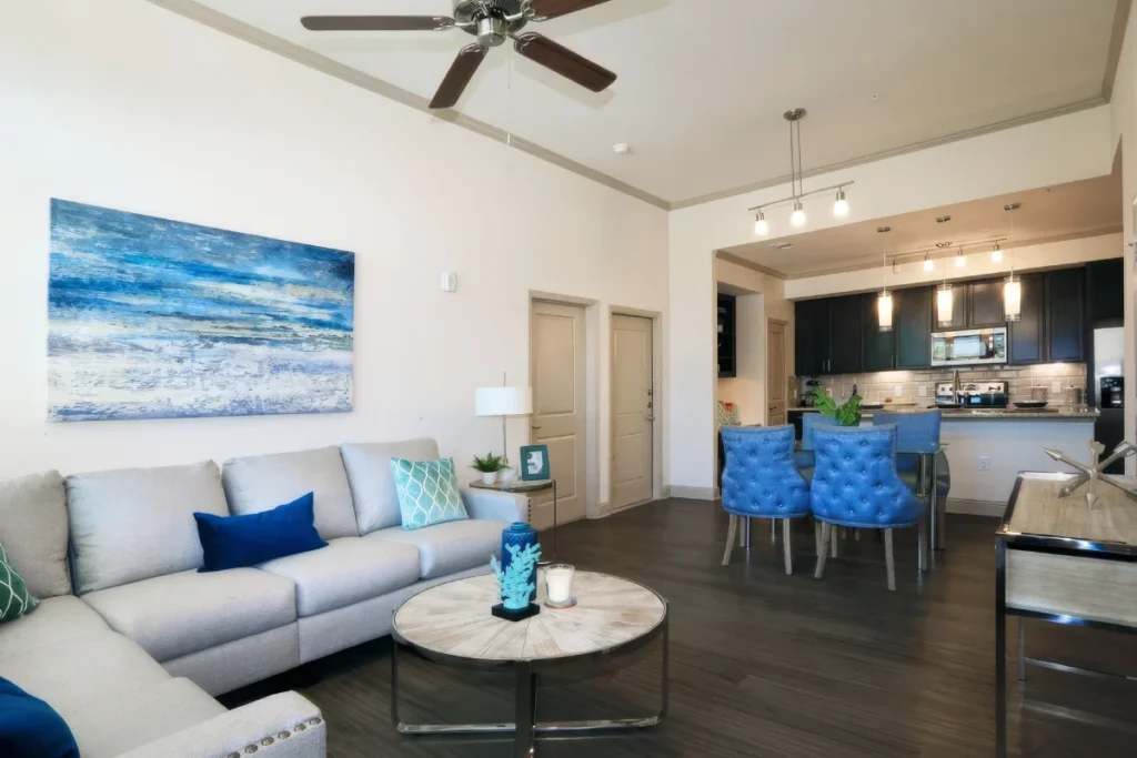 Apartment living area with a light sofa and coastal wall art, with a kitchen island and blue chairs beyond