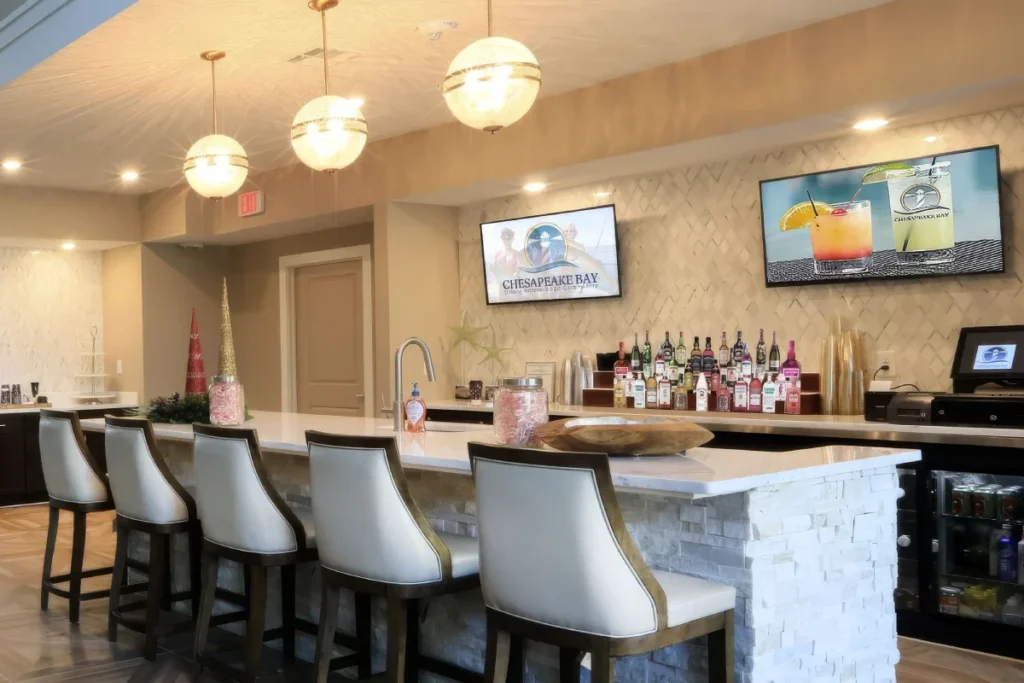 Bar counter with white bar stools, a stone-look front, bottle shelves and wall-mounted TVs