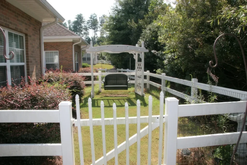 White fence with an arched garden gate beside a brick home and landscaped yard