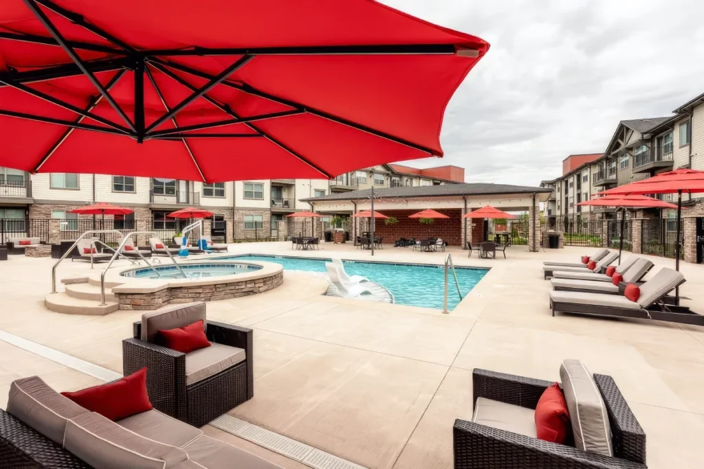 Poolside patio with a red umbrella, lounge chairs and a hot tub, surrounded by apartment buildings