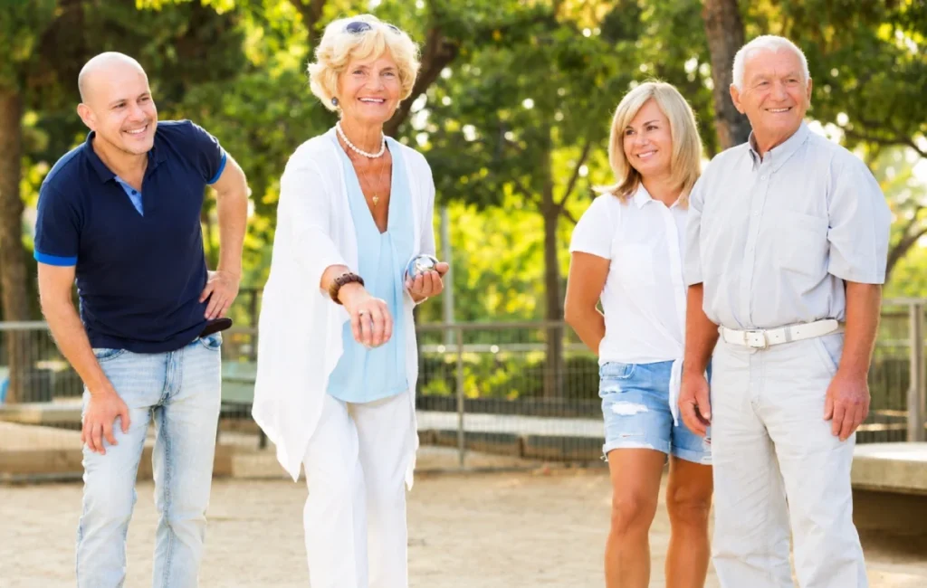 Group of older adults standing on a gravel court watching a game, smiling and chatting