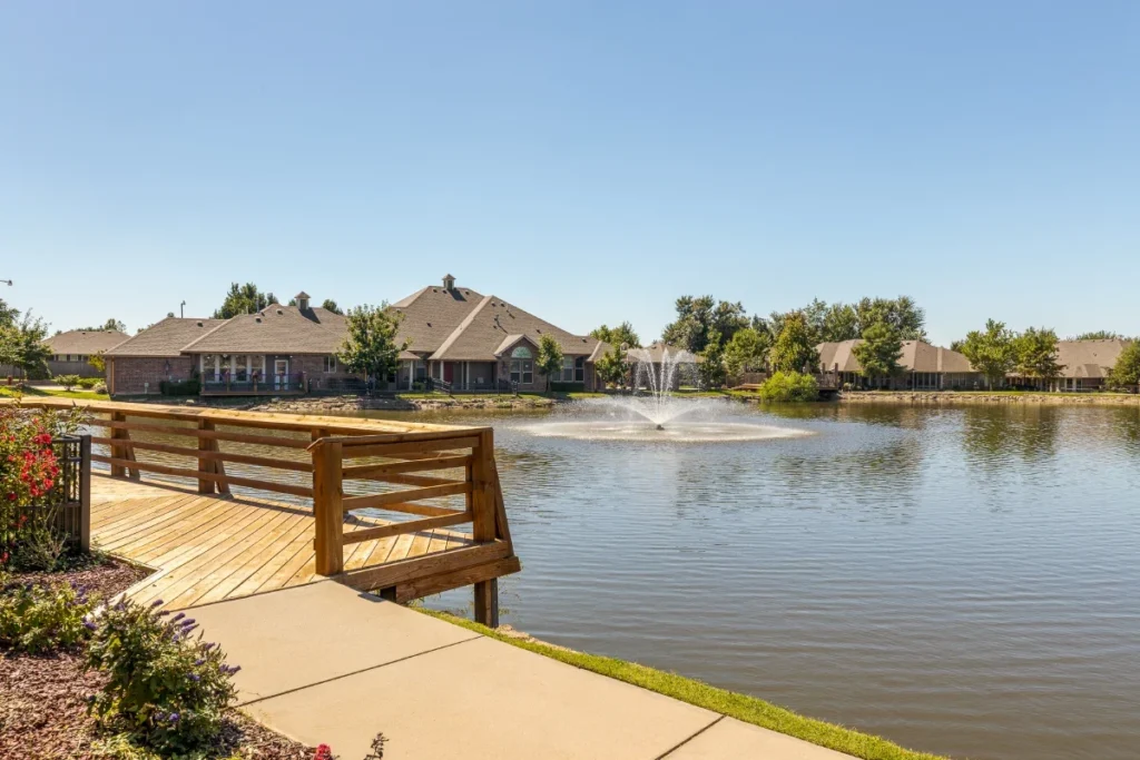 Wooden walkway leading to a pond with a fountain, with homes across the water
